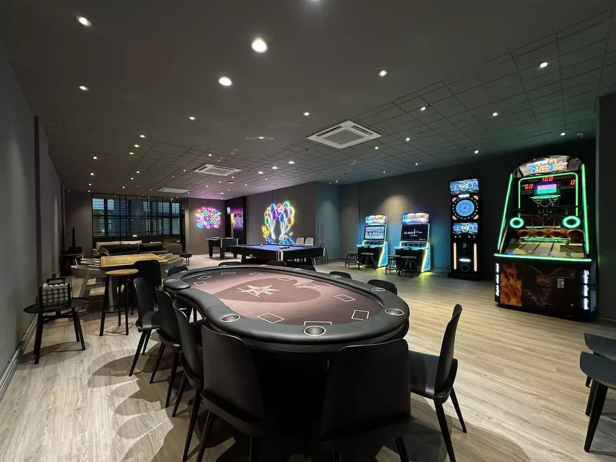 A vibrant entertainment room featuring a central poker table, pool table, and various arcade games under cool-toned lighting.