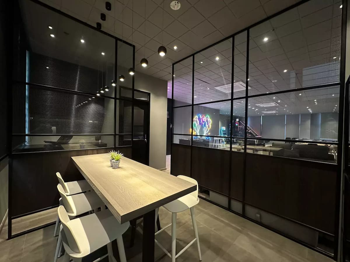 A minimalist workspace features a wooden table and white chairs, separated by a glass and black metal partition from a vibrant social area.