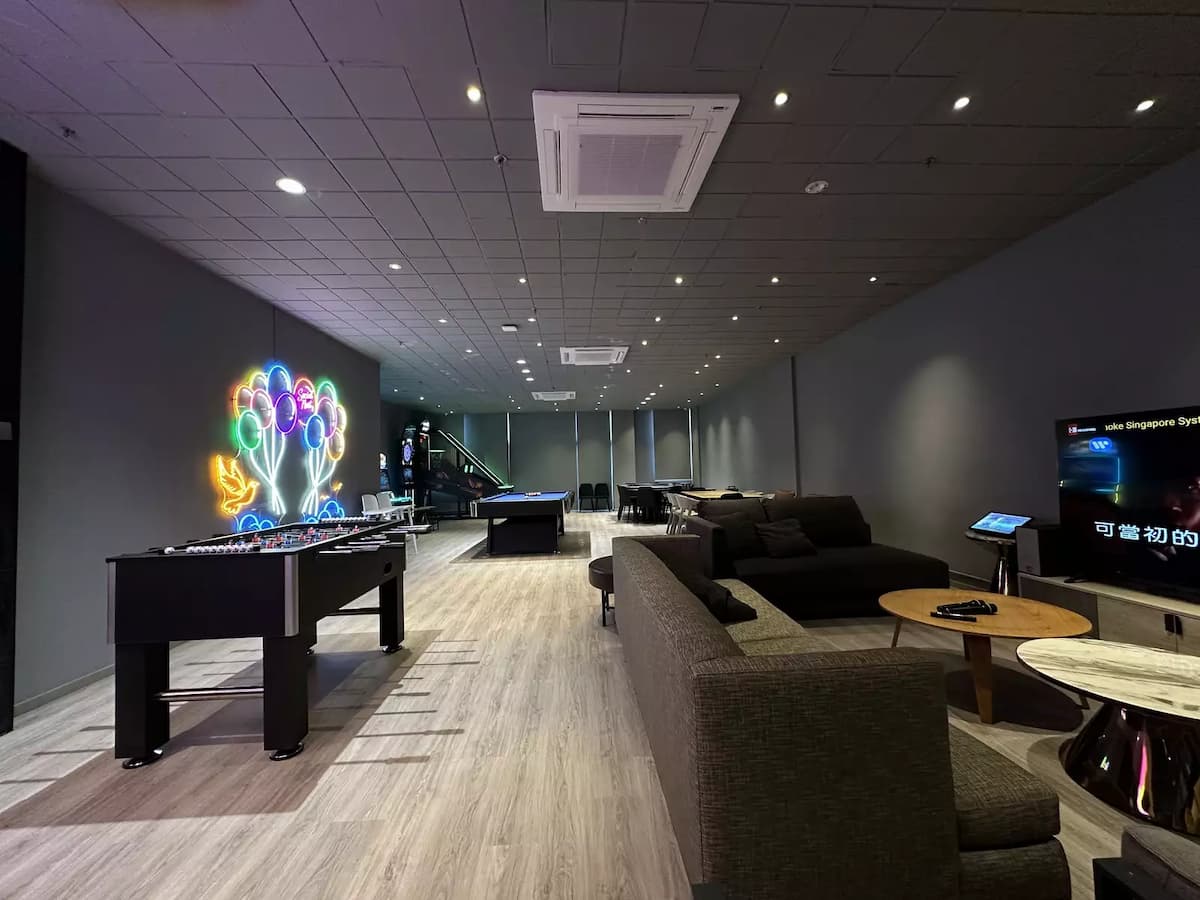 A spacious entertainment lounge with grey sofas, coffee tables, a foosball table, and a colorful neon wall sign under a grid ceiling.