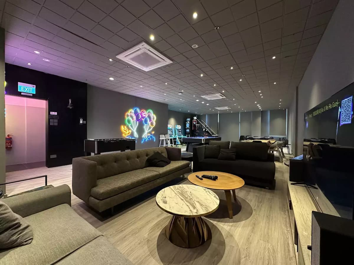 A modern entertainment lounge featuring plush grey sofas, a large TV, and a foosball table under soft, dim lighting with a colorful neon wall sign.