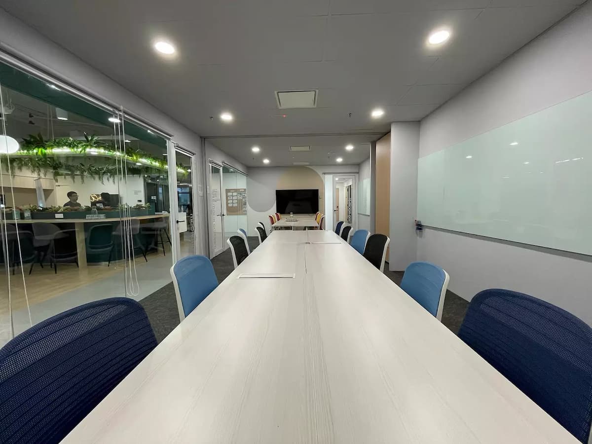 Modern boardroom with long white conference table, mixed blue and black chairs, glass whiteboard, and wall-mounted presentation screen.
