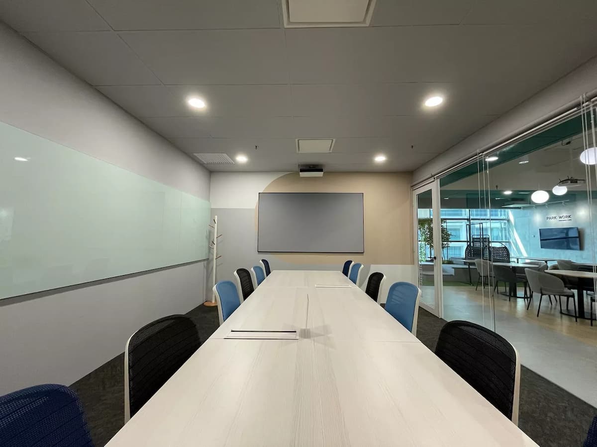 Modern boardroom with long white conference table, mixed blue and black chairs, glass whiteboard, and wall-mounted presentation screen.