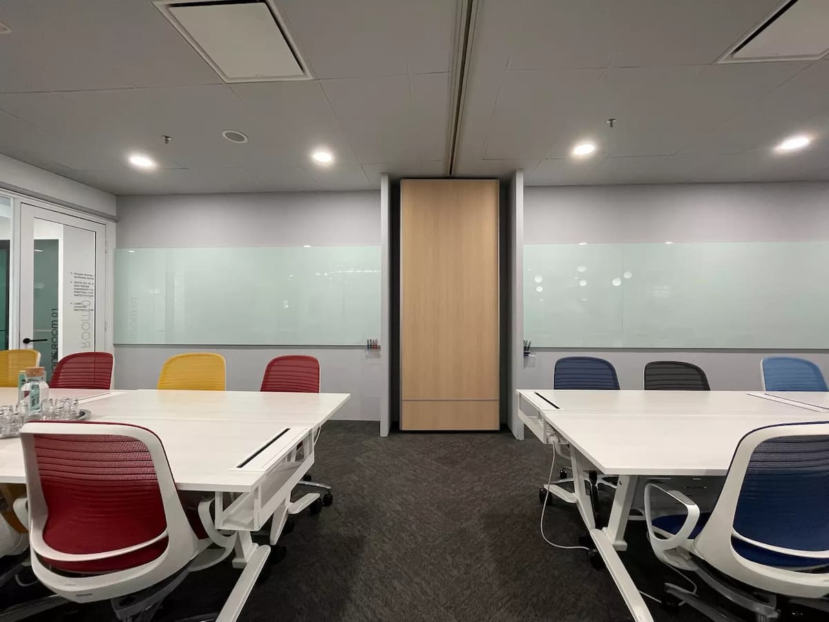 Flexible meeting space with modular white tables, red, yellow, and blue chairs, glass whiteboards, and sliding partition wall.