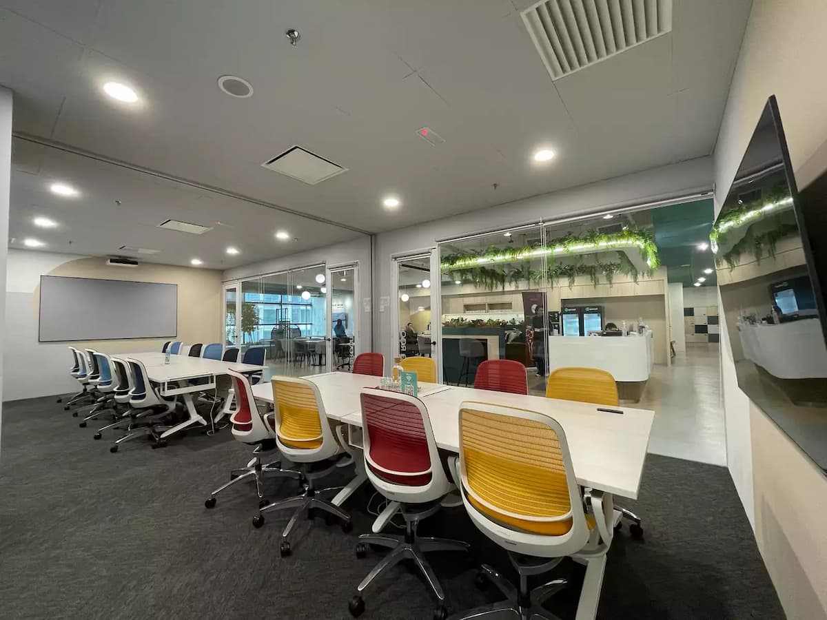 Spacious meeting room with white tables, red and yellow chairs, glass walls, and view into a modern reception area with greenery.