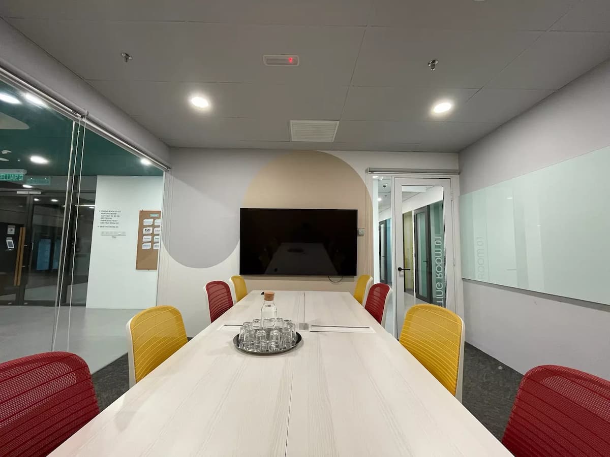 Modern meeting room with long wooden table, red and yellow chairs, wall-mounted TV, and glass partitions on both sides.