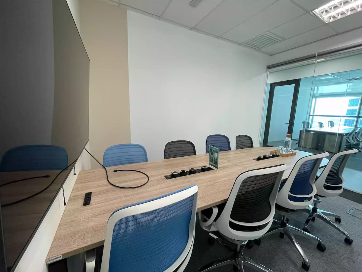 Compact meeting room with oval wooden table, ergonomic chairs, wall-mounted TV, and neutral-toned walls under recessed lighting.
