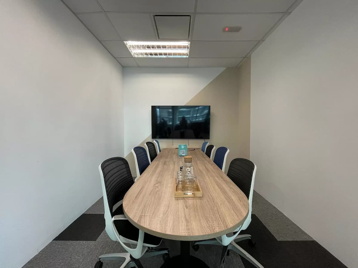 Compact meeting room with oval wooden table, ergonomic chairs, wall-mounted TV, and neutral-toned walls under recessed lighting.