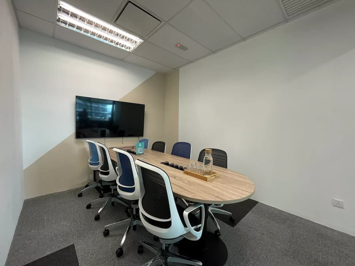 Compact meeting room with oval wooden table, ergonomic chairs, wall-mounted TV, and neutral-toned walls under recessed lighting.