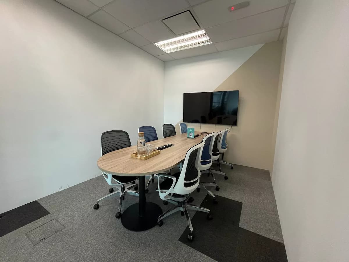 Compact meeting room with oval wooden table, ergonomic chairs, wall-mounted TV, and neutral-toned walls under recessed lighting.
