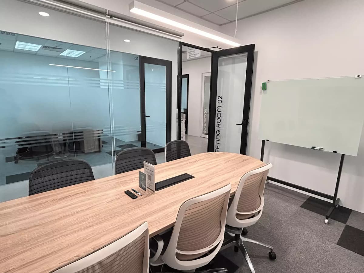 A small meeting room with a wooden conference table, ergonomic chairs, a mobile glass whiteboard, and glass partitions leading to adjacent rooms.