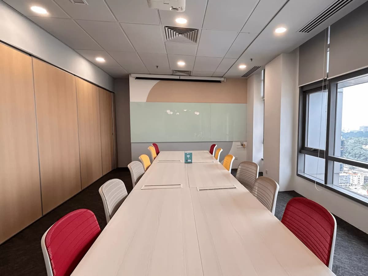 A contemporary boardroom with a long conference table, colorful ergonomic chairs, wood-paneled walls, a glass writing board, and a ceiling-mounted projector.
