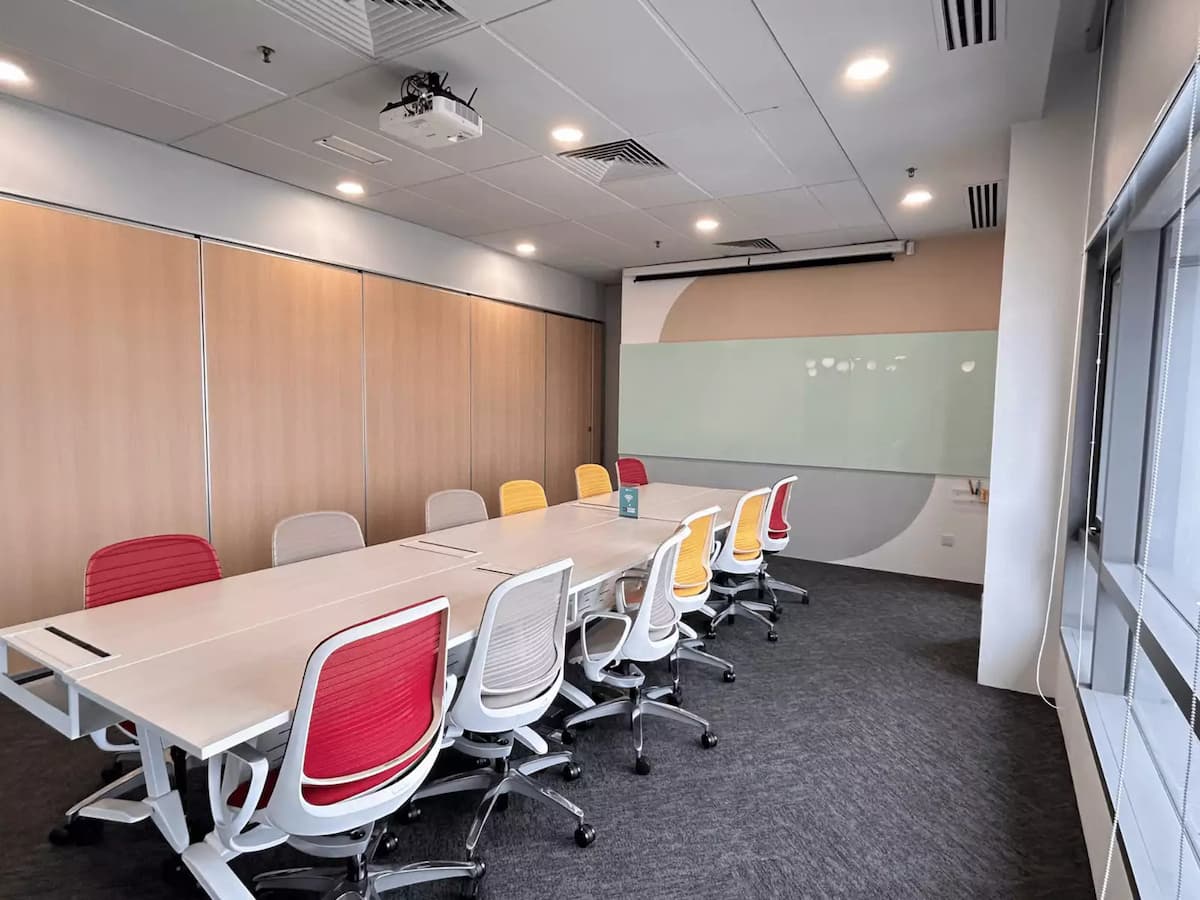 A contemporary boardroom with a long conference table, colorful ergonomic chairs, wood-paneled walls, a glass writing board, and a ceiling-mounted projector.