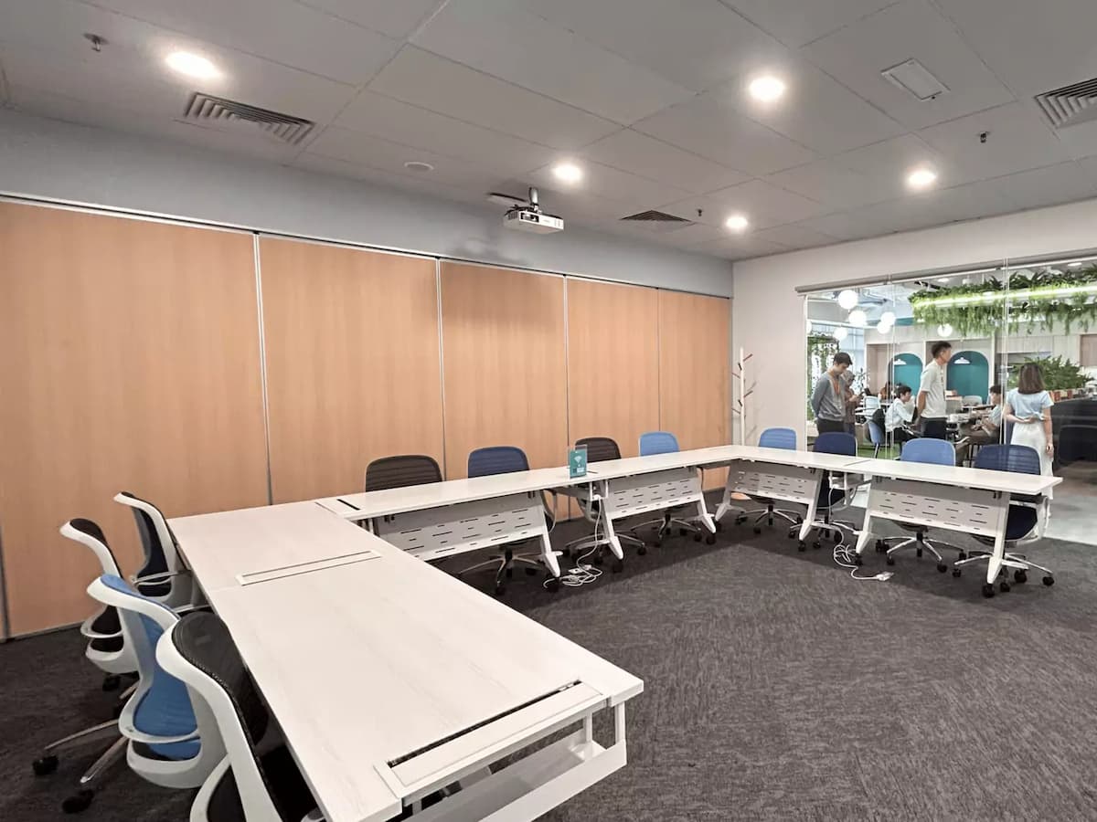 A modern training room with modular white tables arranged in a U-shape, ergonomic chairs, a wall-length glass whiteboard, and a ceiling-mounted projector for presentations.
