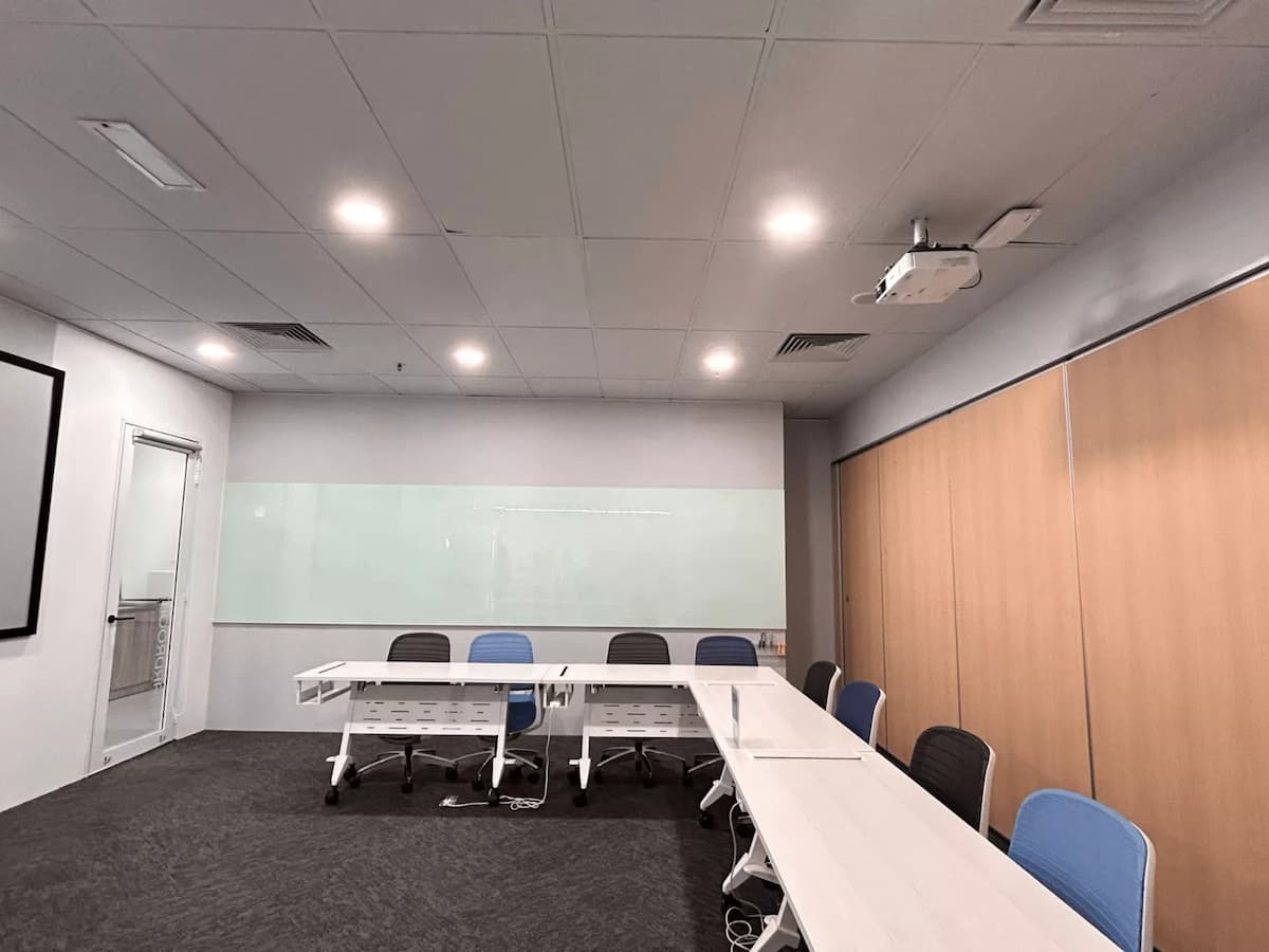 A modern training room with modular white tables arranged in a U-shape, ergonomic chairs, a wall-length glass whiteboard, and a ceiling-mounted projector for presentations.