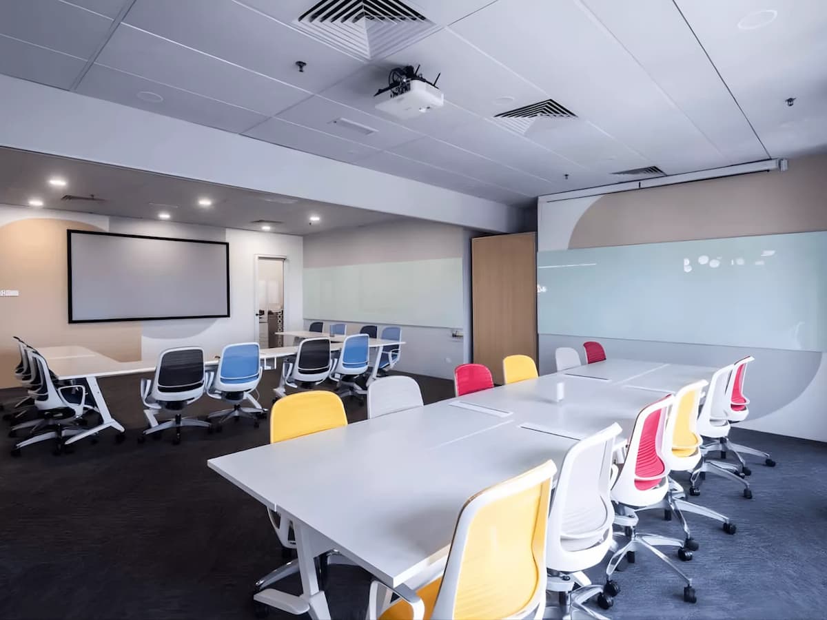 A spacious, modern training room with modular white tables, colorful ergonomic chairs, a large projection screen, and wall-length glass whiteboards—designed for workshops and collaborative sessions.
