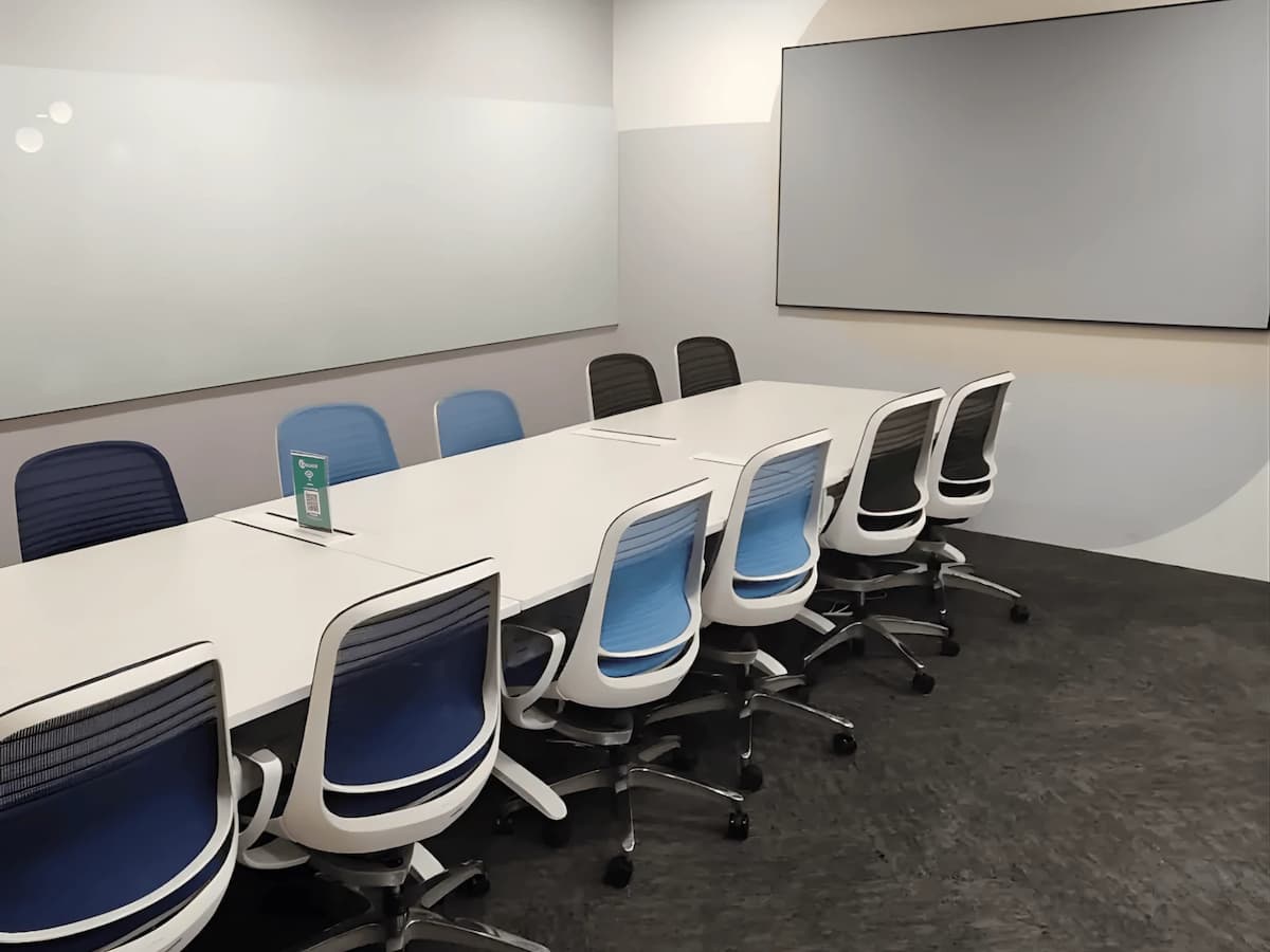 A modern training and meeting room with a long conference table, blue and black ergonomic chairs, a wall-length glass whiteboard, and a large display screen at the front.