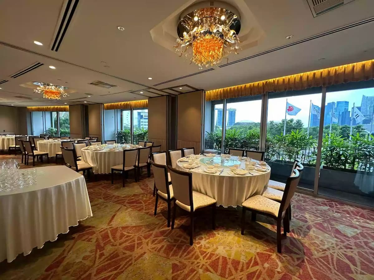 Bright banquet dining space with round tables, elegant chandeliers, patterned carpet, and city views through large windows.