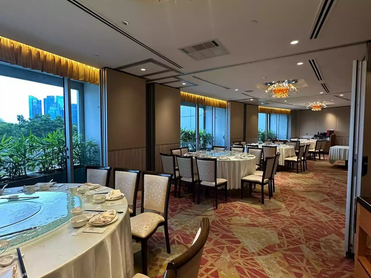 Spacious banquet hall with round tables, refined place settings, warm chandelier lighting, and greenery-lined windows.
