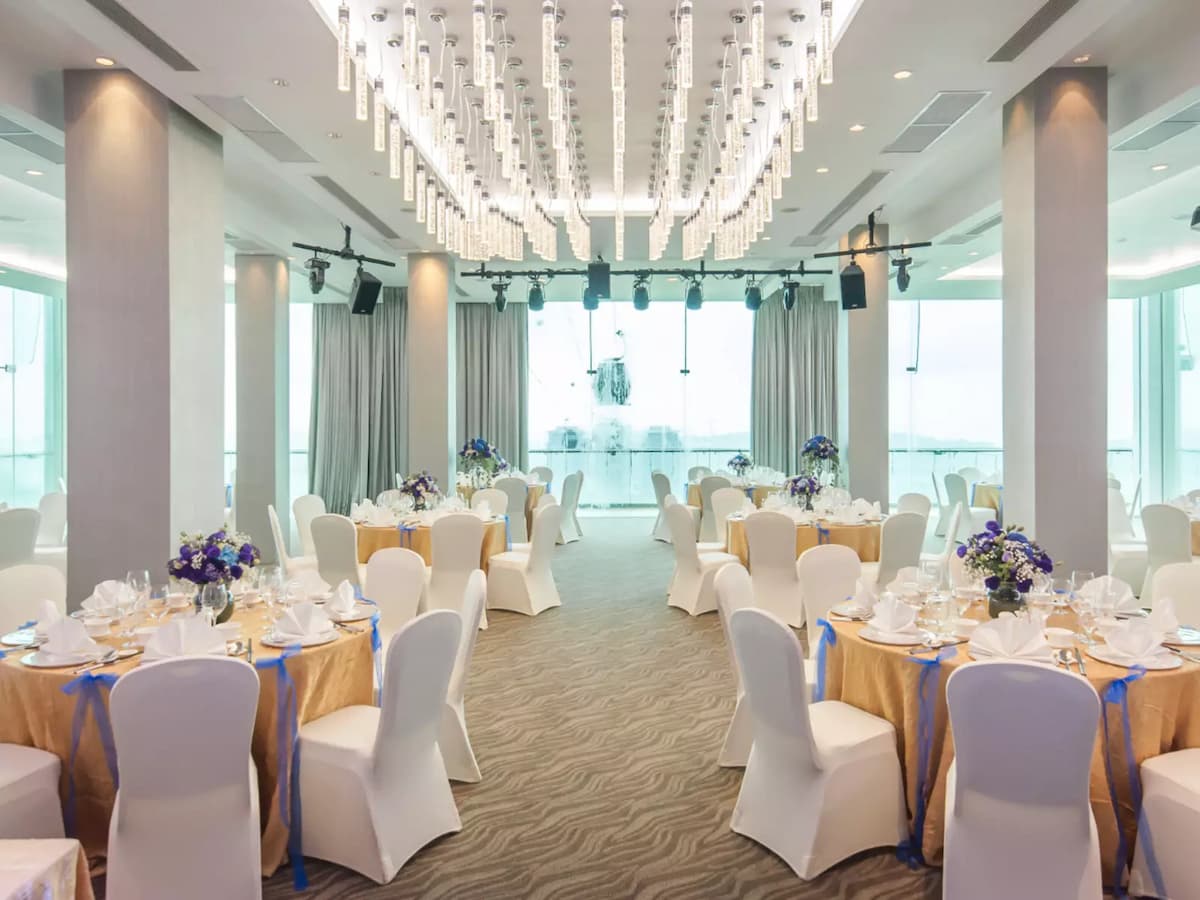 Elegant banquet hall with round tables set for an event, white chair covers, blue-accented table linens, floral centerpieces, and a crystal chandelier.