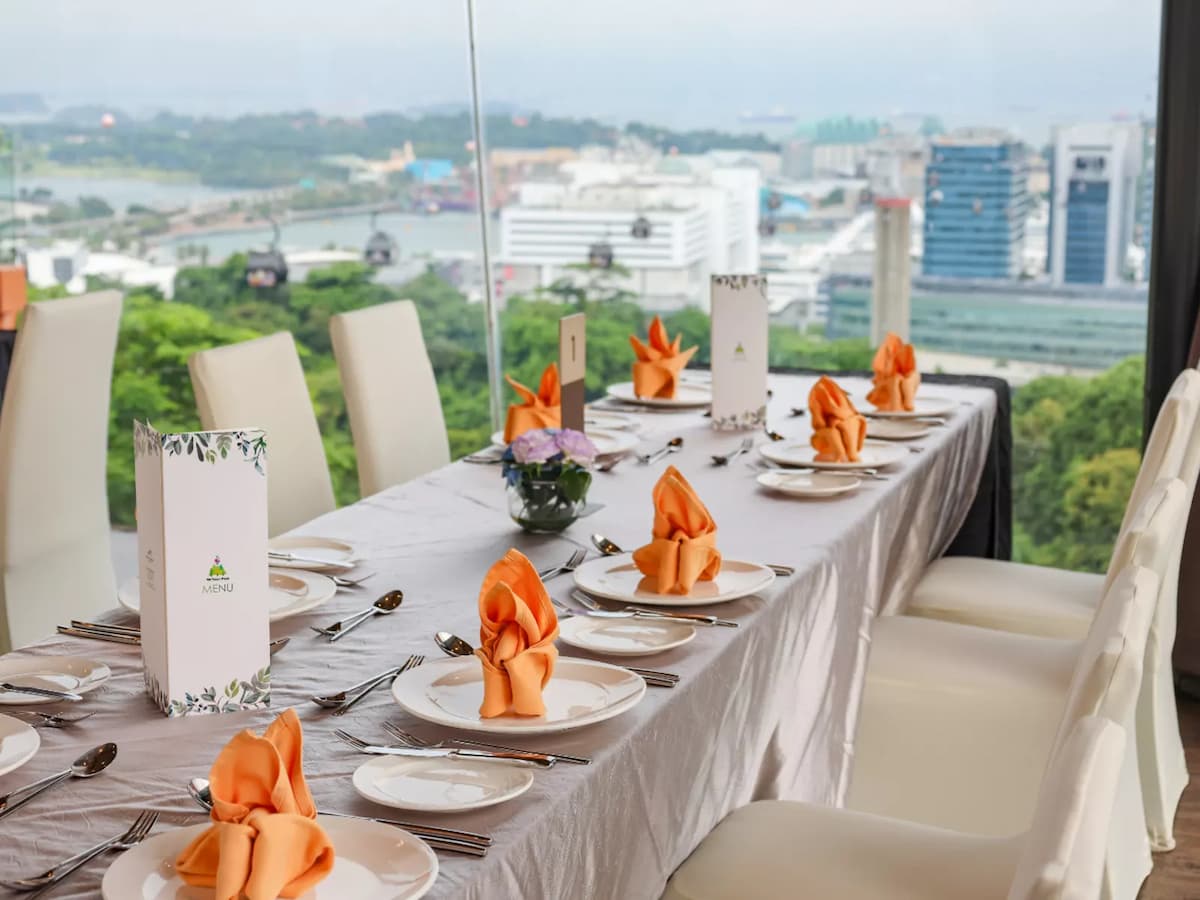 Long banquet table with white tablecloth, orange folded napkins, menu cards, and place settings by a window overlooking city and greenery views.
