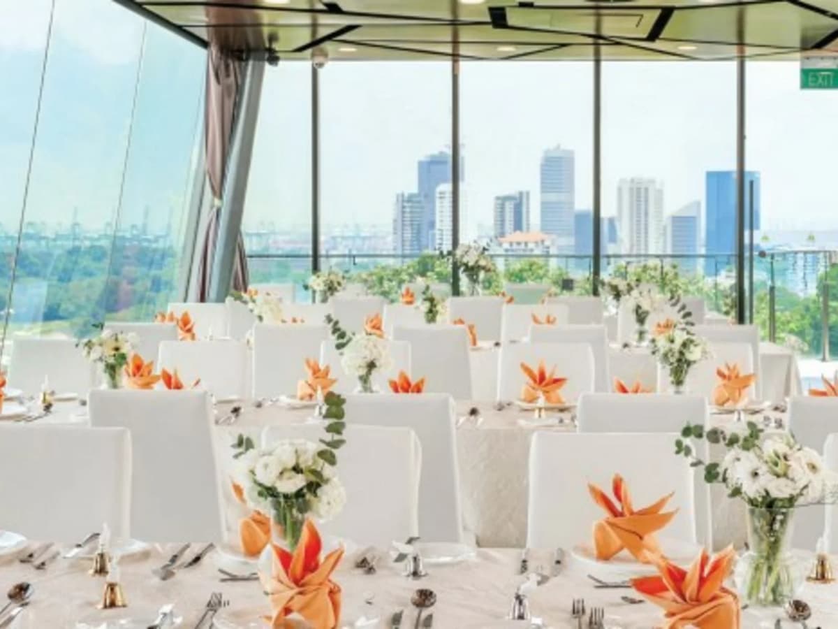 Bright banquet hall with white tables and chairs, orange napkin folds, floral centerpieces, and city skyline views through glass windows.