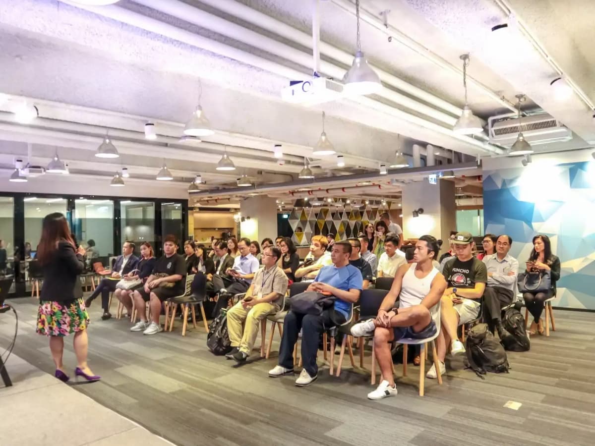 Speaker presenting to seated audience in a modern seminar room with glass walls and geometric wall design.