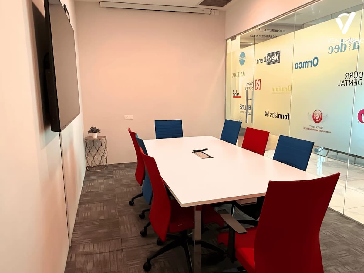 Small meeting room at Mutiara Event Space with boardroom table, colourful chairs, and wall-mounted TV for meetings