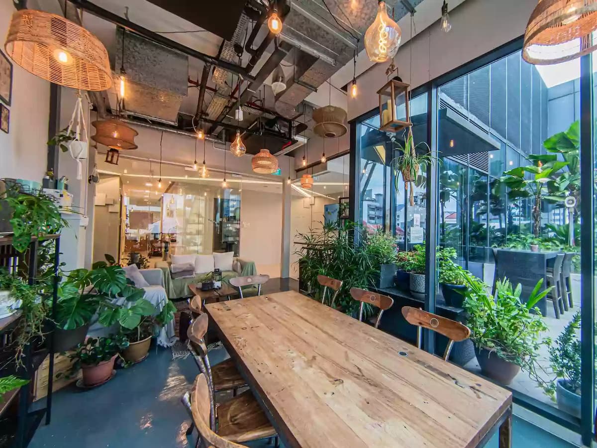 Bright plant-filled meeting space with a communal wooden table and floor-to-ceiling windows overlooking the city—ideal for workshops, team meetings, creative sessions, and small group gatherings.