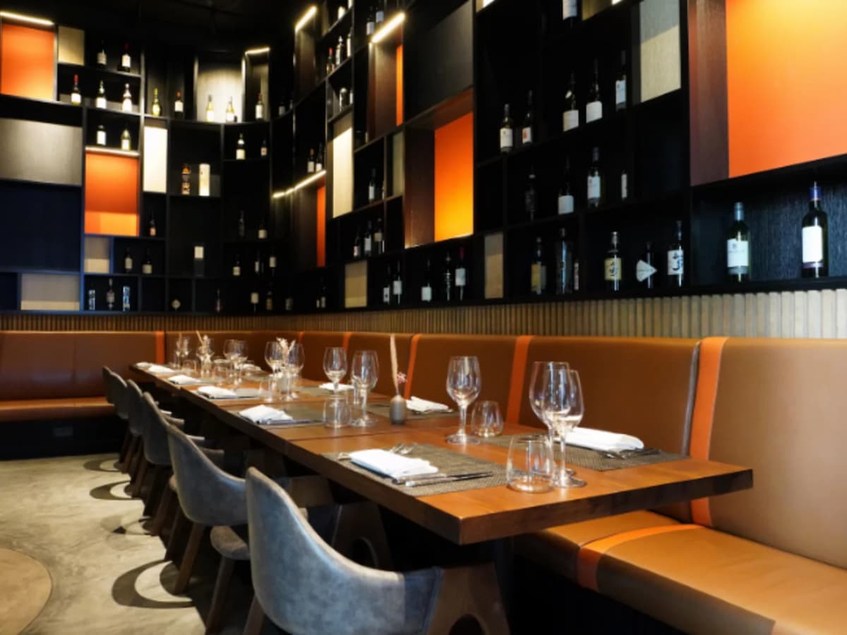 Elegant restaurant seating with long wooden table, leather banquette, wine bottle display wall, and warm accent lighting.