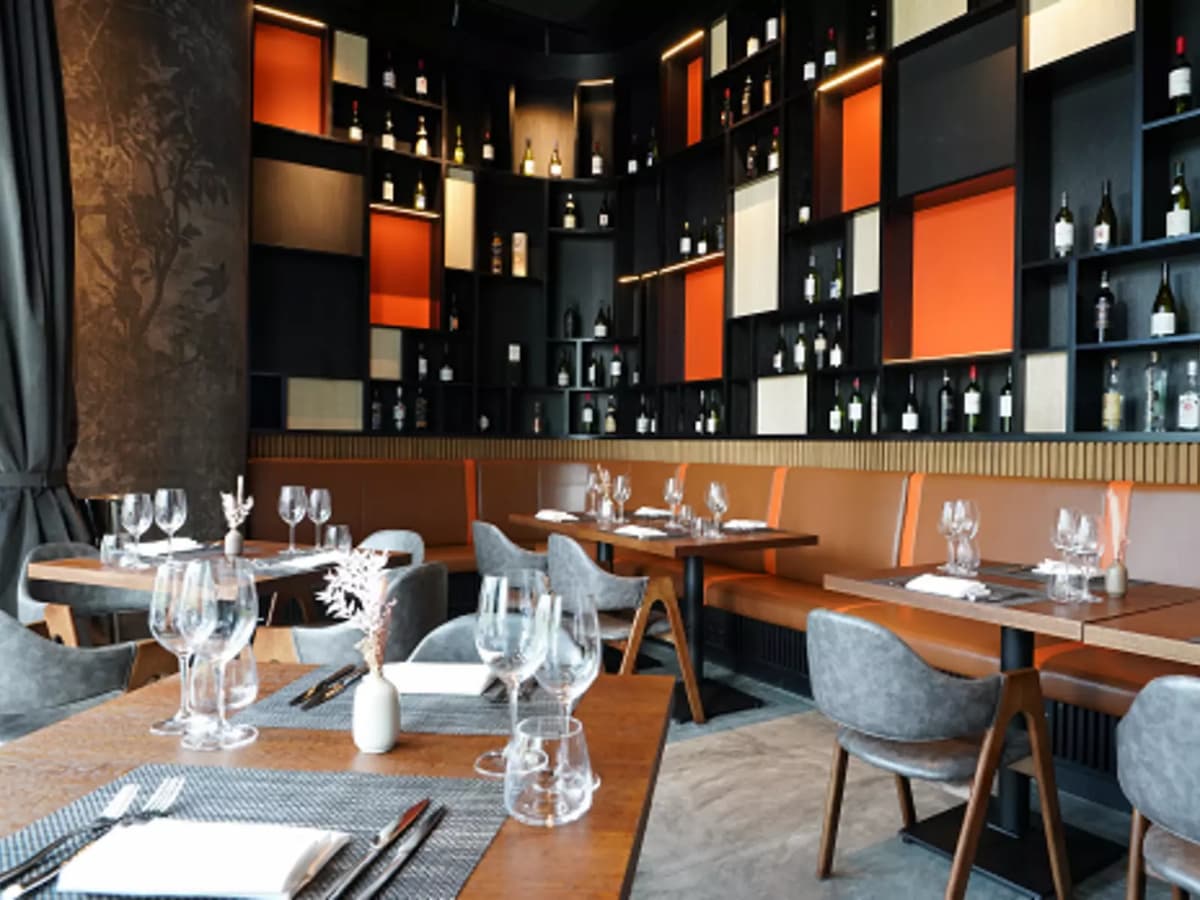 Modern restaurant with wooden tables, grey upholstered chairs, and a feature wall of wine bottles in black and orange shelving.Modern restaurant with wooden tables, grey upholstered chairs, and a feature wall of wine bottles in black and orange shelving.