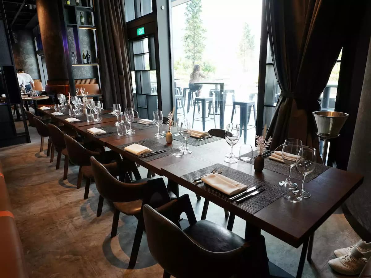 Refined restaurant dining area with long wooden table set with glassware and napkins, beside tall windows and balcony seating.