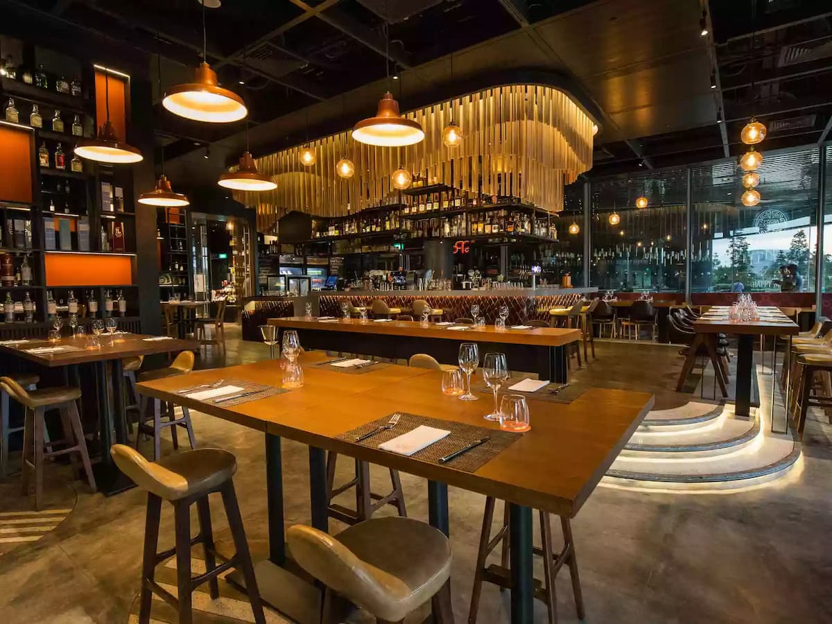 Upscale restaurant interior with wooden tables, leather chairs, warm pendant lighting, and a stylish open kitchen backdrop.